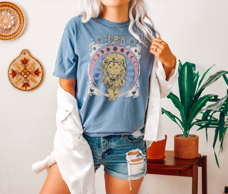 May include: A blue t-shirt featuring a graphic of a lion and the word "Leo" in a retro style. The shirt is being worn by a person with a white denim jacket and denim shorts.