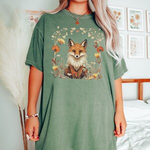 以下が含まれることがあります： 花で囲まれた狐が描かれた緑色のオーバーサイズTシャツ。狐はオレンジと白で、目は黒です。