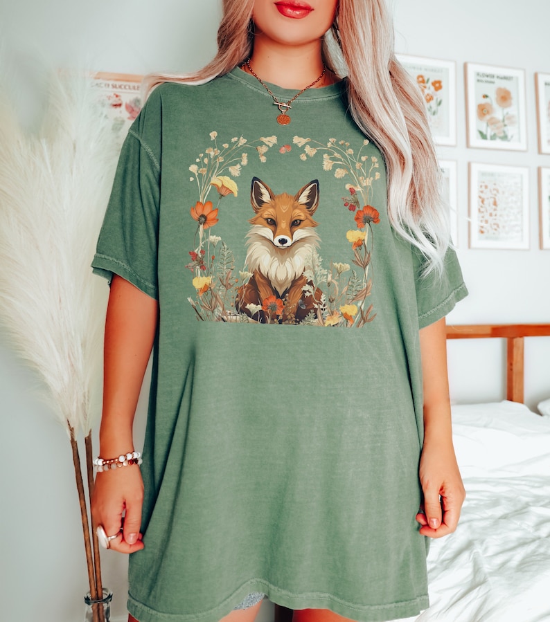 以下が含まれることがあります： 花で囲まれた狐が描かれた緑色のオーバーサイズTシャツ。狐はオレンジと白で、目は黒です。