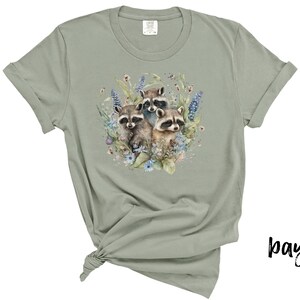 May include: Sage green t-shirt featuring a watercolor illustration of three raccoons surrounded by flowers and foliage. The design includes blue, purple, and pink floral elements. The shirt has rolled-up sleeves and a knotted hem.