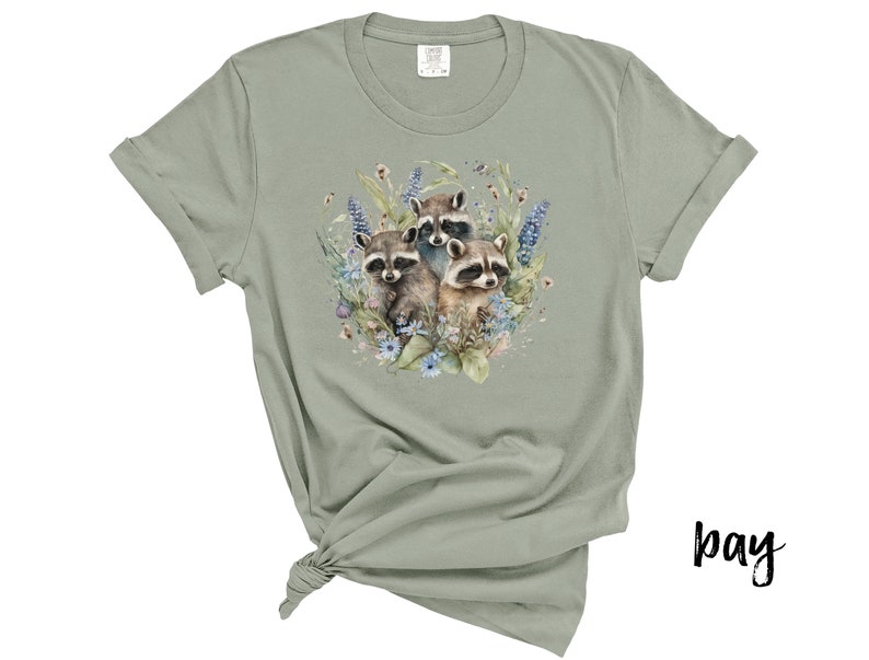 May include: Sage green t-shirt featuring a watercolor illustration of three raccoons surrounded by flowers and foliage. The design includes blue, purple, and pink floral elements. The shirt has rolled-up sleeves and a knotted hem.
