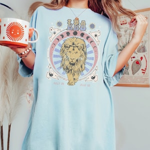 May include: A light blue t-shirt with a vintage-style graphic of a lion with the word "LEO" and the dates "July 23" and "Aug 22" printed on it.