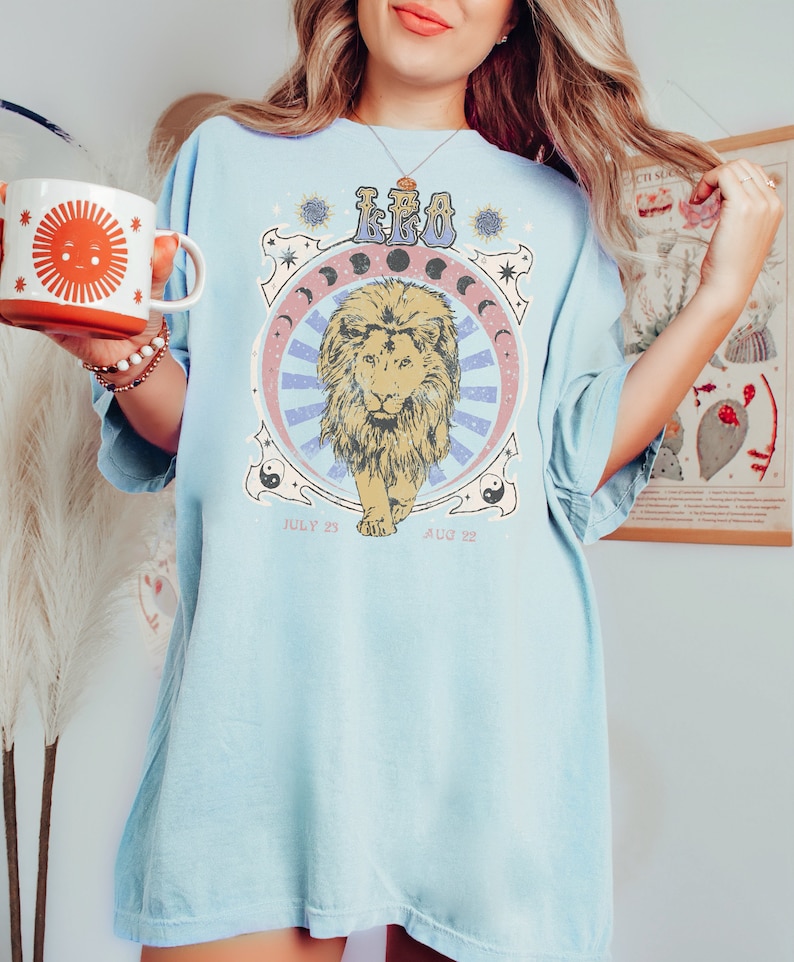 May include: A light blue t-shirt with a vintage-style graphic of a lion with the word "LEO" and the dates "July 23" and "Aug 22" printed on it.