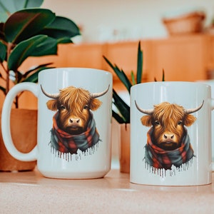 Pode incluir: Duas canecas de cer&acirc;mica branca com um design de vaca das Terras Altas. A vaca tem pelo castanho, chifres grandes e usa um cachecol xadrez vermelho e preto. As canecas est&atilde;o em uma superf&iacute;cie rosa claro.