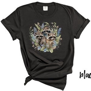 May include: Black t-shirt featuring a watercolor illustration of three raccoons surrounded by blue and green floral elements. The raccoons have brown and gray fur. The shirt is knotted at the bottom.