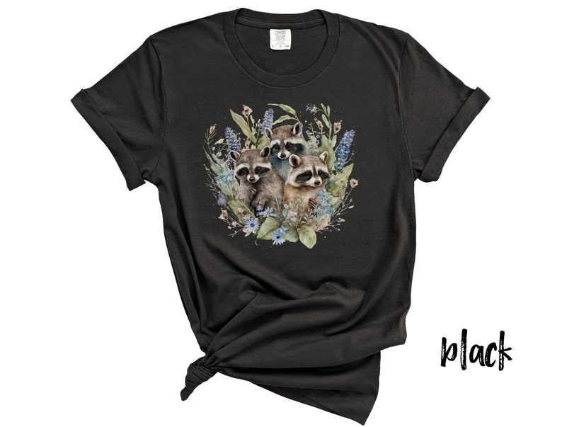 May include: Black t-shirt featuring a watercolor illustration of three raccoons surrounded by blue and green floral elements. The raccoons have brown and gray fur. The shirt is knotted at the bottom.