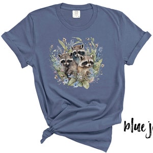 May include: Blue jean colored t-shirt featuring a watercolor illustration of three raccoons surrounded by flowers and foliage. The raccoons have gray and brown fur with black masks. The shirt is made of soft material.