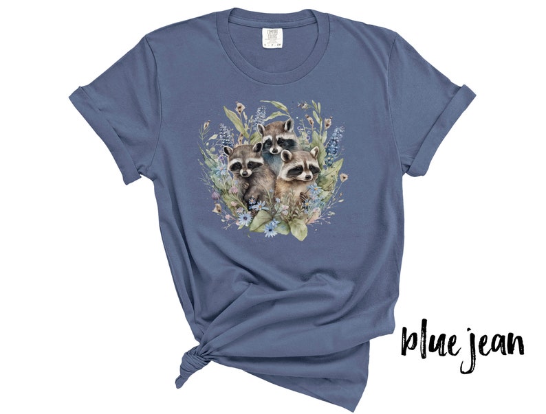 May include: Blue jean colored t-shirt featuring a watercolor illustration of three raccoons surrounded by flowers and foliage. The raccoons have gray and brown fur with black masks. The shirt is made of soft material.
