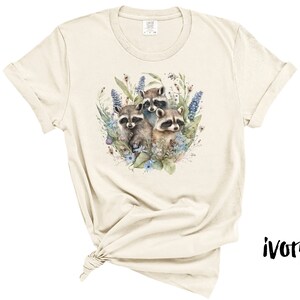 May include: Ivory-colored t-shirt featuring a watercolor illustration of three raccoons surrounded by blue and purple flowers and greenery. The shirt has short sleeves and a crew neck. The word "ivory" is printed in black on the bottom right.