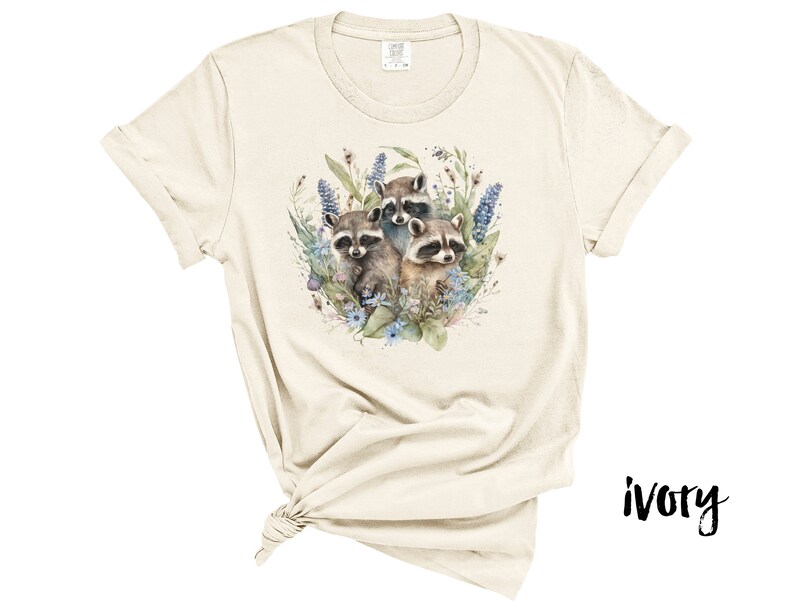 May include: Ivory-colored t-shirt featuring a watercolor illustration of three raccoons surrounded by blue and purple flowers and greenery. The shirt has short sleeves and a crew neck. The word "ivory" is printed in black on the bottom right.