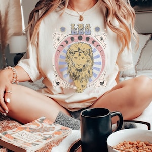 May include: A woman wearing a cream coloured t-shirt with a vintage style graphic of a lion and the text "LEO AUG 22".
