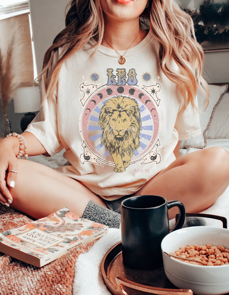 May include: A woman wearing a cream coloured t-shirt with a vintage style graphic of a lion and the text "LEO AUG 22".