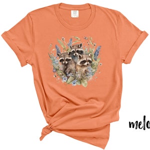 May include: Melon-colored t-shirt featuring a watercolor illustration of three raccoons surrounded by blue and purple flowers and greenery. The shirt has short sleeves and a crew neck. The word "melon" is printed in black script on the bottom right.