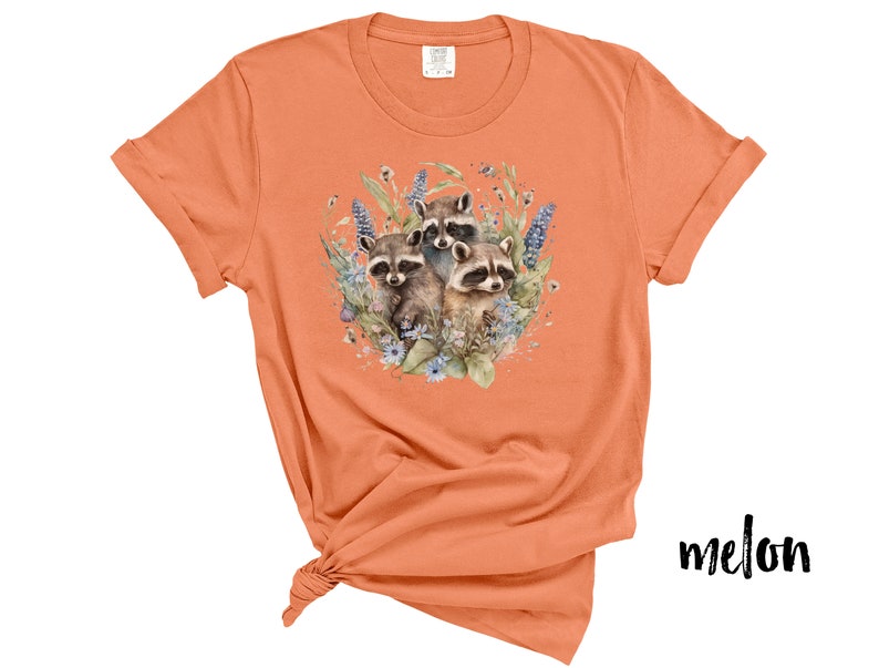 May include: Melon-colored t-shirt featuring a watercolor illustration of three raccoons surrounded by blue and purple flowers and greenery. The shirt has short sleeves and a crew neck. The word "melon" is printed in black script on the bottom right.