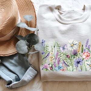 Peut inclure: Un sweat-shirt gris clair avec un motif floral imprimé. L'imprimé présente une variété de fleurs, dont des marguerites, de la lavande et des fleurs sauvages, dans des tons de violet, rose, blanc et jaune. On y trouve également des papillons et des coccinelles.