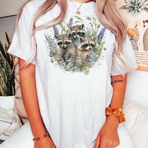 May include: White t-shirt featuring a watercolor illustration of three raccoons surrounded by blue and green foliage. The raccoons have gray and black fur. The shirt is a light color and has short sleeves.