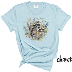May include: Light blue chambray t-shirt featuring a watercolor illustration of three raccoons surrounded by flowers and foliage. The design includes shades of brown, green, and purple. The shirt has rolled-up sleeves and a tied knot at the bottom.