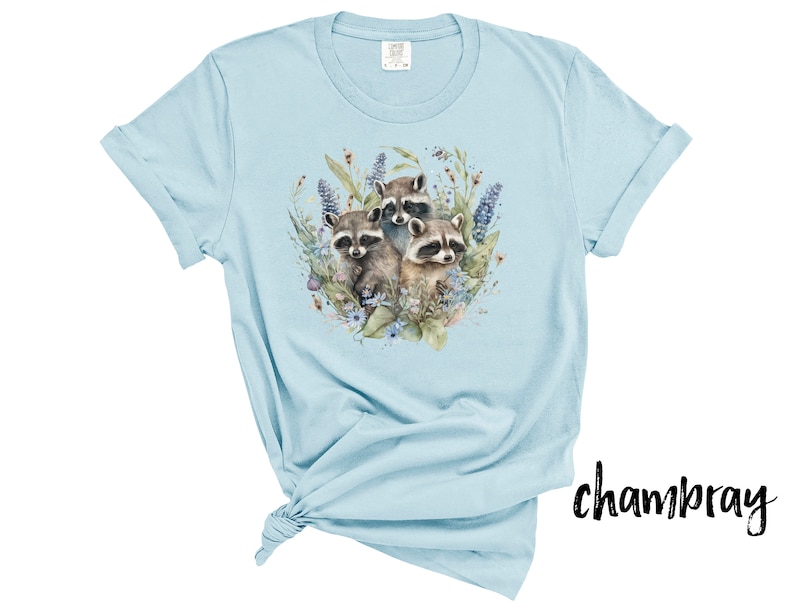 May include: Light blue chambray t-shirt featuring a watercolor illustration of three raccoons surrounded by flowers and foliage. The design includes shades of brown, green, and purple. The shirt has rolled-up sleeves and a tied knot at the bottom.