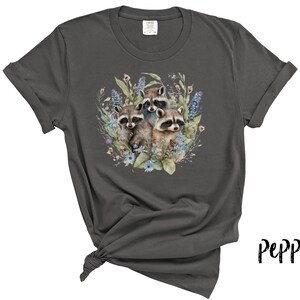May include: Dark grey t-shirt featuring a watercolor illustration of three raccoons surrounded by blue and green flowers and foliage. The shirt has rolled-up sleeves and a knotted hem. The word "Pepper" is written in a script font.