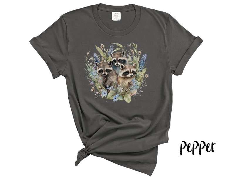 May include: Dark grey t-shirt featuring a watercolor illustration of three raccoons surrounded by blue and green flowers and foliage. The shirt has rolled-up sleeves and a knotted hem. The word "Pepper" is written in a script font.