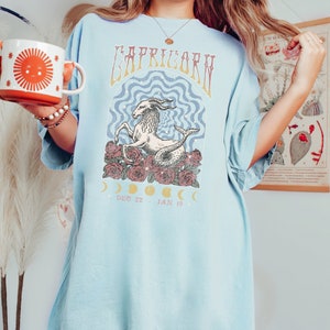 May include: A light blue t-shirt with a graphic of a Capricorn zodiac sign with a sea-goat, roses, and moons. The text "Capricorn" is printed in a vintage style font. The dates "Dec 22 - Jan 19" are printed below the graphic.