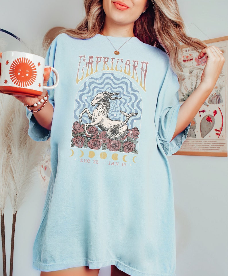 May include: A light blue t-shirt with a graphic of a Capricorn zodiac sign with a sea-goat, roses, and moons. The text "Capricorn" is printed in a vintage style font. The dates "Dec 22 - Jan 19" are printed below the graphic.