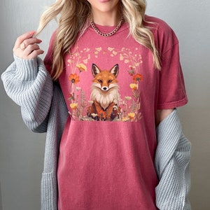 以下が含まれることがあります： 花に囲まれたキツネのグラフィックが描かれた赤いTシャツ。キツネはオレンジと白で、目は黒く、鼻は黒いです。