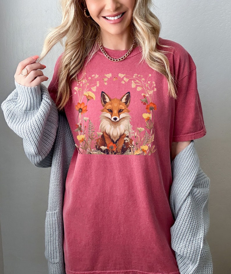 以下が含まれることがあります： 花に囲まれたキツネのグラフィックが描かれた赤いTシャツ。キツネはオレンジと白で、目は黒く、鼻は黒いです。