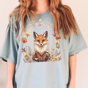 以下が含まれることがあります： 花で囲まれた狐が描かれた薄い青色のTシャツ。狐はオレンジと白で、黒い鼻と目です。