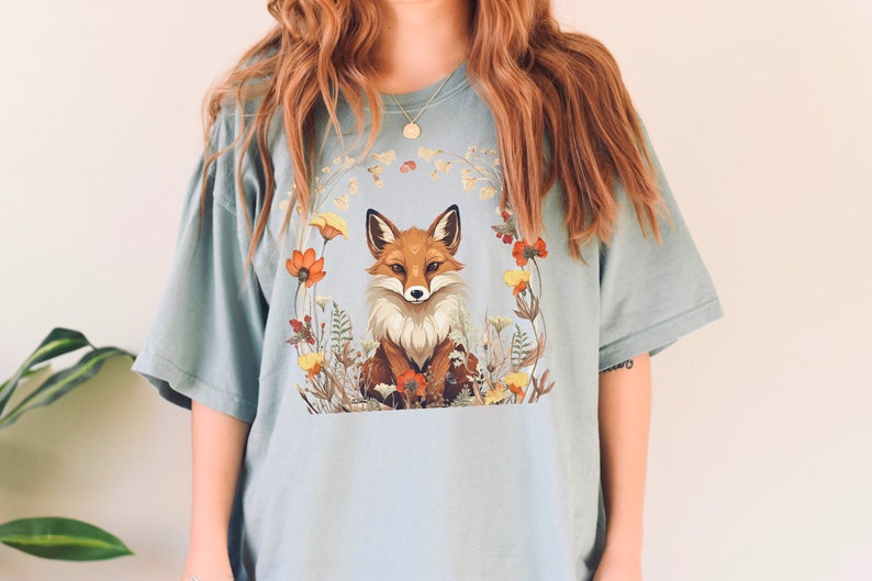 以下が含まれることがあります： 花で囲まれた狐が描かれた薄い青色のTシャツ。狐はオレンジと白で、黒い鼻と目です。
