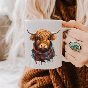 Pode incluir: Caneca de cer&acirc;mica branca com uma ilustra&ccedil;&atilde;o em aquarela de uma vaca das Terras Altas. A vaca tem pelo e chifres castanhos e usa um cachecol xadrez vermelho, azul e verde. A caneca &eacute; segurada por uma pessoa.