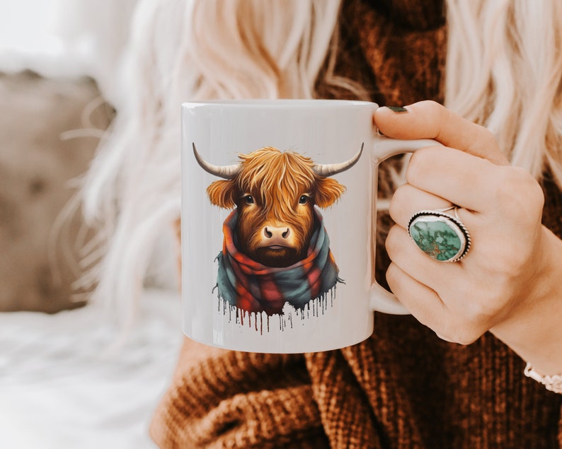Pode incluir: Caneca de cer&acirc;mica branca com uma ilustra&ccedil;&atilde;o em aquarela de uma vaca das Terras Altas. A vaca tem pelo e chifres castanhos e usa um cachecol xadrez vermelho, azul e verde. A caneca &eacute; segurada por uma pessoa.