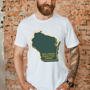 May include: White t-shirt with a green outline of the state of Wisconsin and the text "We'll never forget you, Daron!"