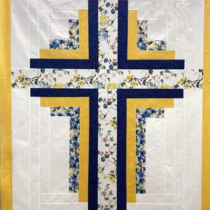 May include: A handmade quilt with a cross design. The cross is made with white, yellow, and navy blue fabric. Floral patterned fabric is used in the center and in some sections of the cross. The quilt has a yellow border. The quilt is likely made of cotton.