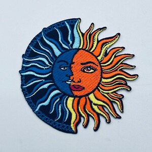 Könnte beinhalten: Gestickter Patch mit einem Sonnen- und Monddesign. Die Sonne ist orange und gelb mit einem menschlichen Gesicht, während der Mond blau und weiß mit einem menschlichen Gesicht ist. Sonne und Mond sind in einer Halbmondform miteinander verbunden.