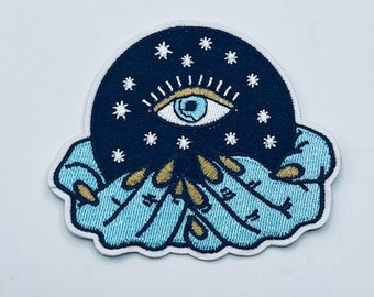 Puede incluir: Un parche bordado azul y negro con un ojo estilizado rodeado de estrellas, sostenido por dos manos con detalles dorados.