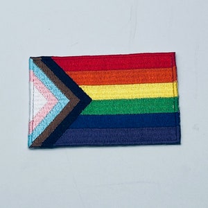 Op de afbeelding: Een regenboog-pride-vlag met een bruine, zwarte, lichtblauwe, roze en witte driehoek in de linkerbovenhoek. De vlag is gemaakt van stof en heeft een gestikte rand.