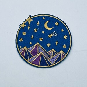 May include: Embroidered patch featuring a night sky design. The patch has a navy blue background with gold stars, a crescent moon, and a shooting star. The bottom of the patch features purple and gold mountains.