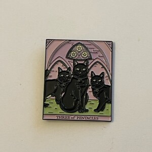 Könnte beinhalten: Quadratischer Ansteckpin mit drei schwarzen Katzen in einem Tarotkarten-Design. Der Pin hat einen rosa-violetten Hintergrund mit dem Text "THREE OF PENTACLES" unten. Die Katzen befinden sich auf einer grünen Oberfläche.
