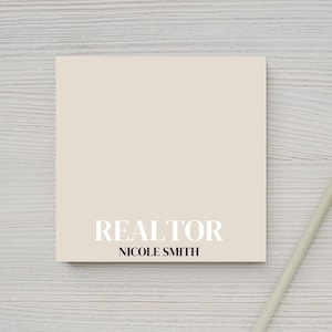 Puede incluir: Un bloc de notas beige con el texto "REALTOR" y "NICOLE SMITH" impreso en negro en la parte delantera. Un lápiz blanco está sobre una superficie de madera blanca junto al bloc de notas.