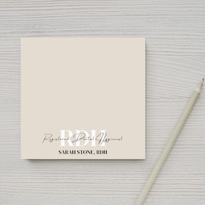 May include: A beige notepad with the text "R.D.H. Registered Dental Hygienist SARAH STONE, R.D.H." printed on it. A pencil is lying on a white wooden surface next to the notepad.
