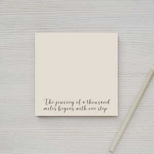 May include: A beige notepad with the quote "The journey of a thousand miles begins with one step" printed on it. A pencil is lying on a white wooden surface next to the notepad.