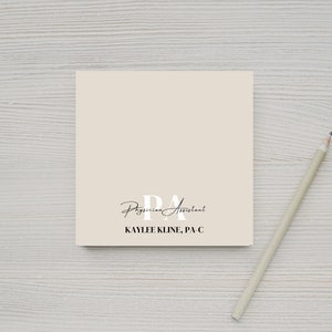 May include: A beige notepad with the text "PA Physician Assistant KAYLEE KLINE, PA-C" printed on it. A pencil is lying on a white wooden surface next to the notepad.