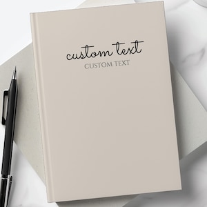 Peut inclure: Un carnet beige avec le texte "custom text" écrit en cursive noire sur la couverture. Le carnet est posé sur une surface grise.