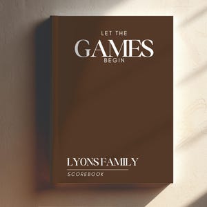 Puede incluir: Un libro de registro marrón con el texto "LET THE GAMES BEGIN" en blanco. Debajo del texto está "LYONS FAMILY SCOREBOOK" en blanco. El libro es rectangular y tiene una sombra en el lado izquierdo.