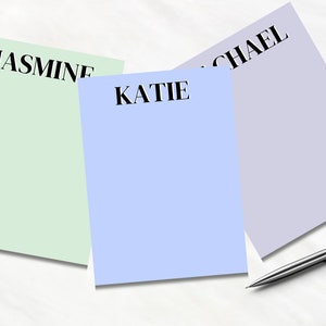 May include: Three blank notepads with different colored backgrounds. The notepads are labeled "Jasmine", "Katie", and "Michael". A silver pen is lying on the white surface.