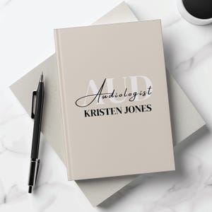May include: Beige notebook with the text "Audiologist" in black script and "KRISTEN JONES" in black block letters.
