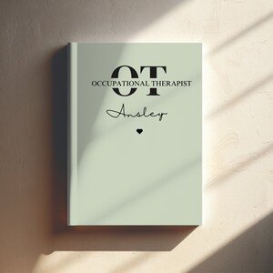 Puede incluir: Una libreta verde claro con el texto "OT OCCUPATIONAL THERAPIST" y el nombre "Ansley" escrito en cursiva. Un pequeño corazón negro está debajo del nombre.