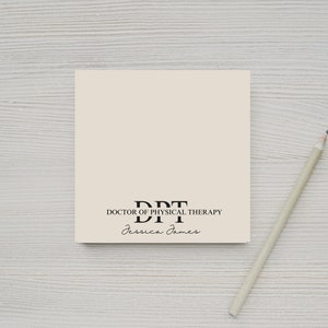 May include: A square, cream-colored notepad with the text "DPT DOCTOR OF PHYSICAL THERAPY Jessica James" in black. A light gray pencil rests diagonally across a light wood-grain surface.
