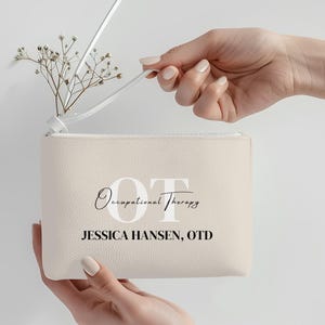 Peut inclure: Une pochette beige à fermeture éclair avec le texte "OT Occupational Therapy" et "JESSICA HANSEN, OTD" imprimé sur le devant. La pochette est tenue par une personne, et une petite brindille de fleurs séchées est à l'intérieur. La pochette a une sangle blanche.